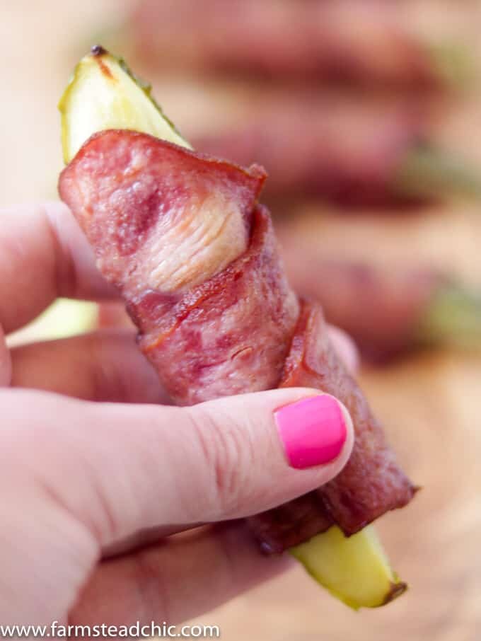 Bacon-wrapped pickles on a natural wood cutting board held by a woman with hot pink gel fingernails