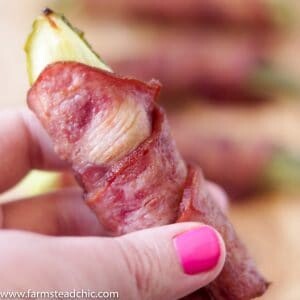 Bacon-wrapped pickles on a natural wood cutting board held by a woman with hot pink gel fingernails