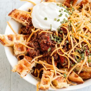 Chow down on these Low Carb, Keto Chili Cheese Chaffle Fries when you're craving some real comfort food. Perfectly firm and crispy chaffles are sliced into "fries," piled with meaty, beanless chili and topped with cheddar cheese, a hefty dollop of sour cream and a sprinkling of vibrant, flavorful chives.